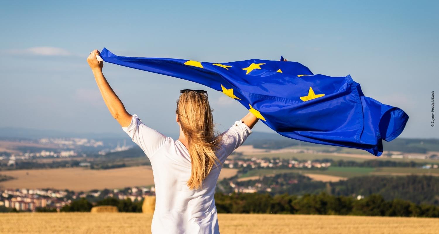 Eine blonde Frau hält eine wehende EU-Fahne in beiden Händen und steht dabei mit dem Rücken zu uns. Sie steht in einem Feld. 