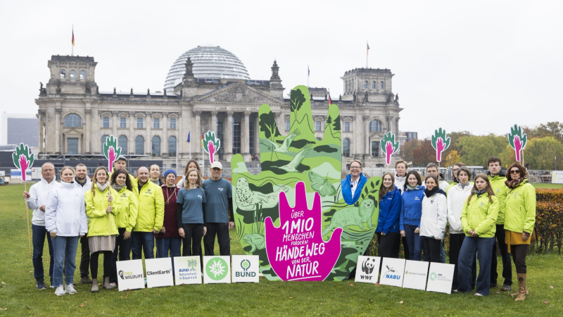 Vertreter:innen verschiedener Umwelt- und Naturschutzorganisationen stehen vor dem Reichstagsgebäude in Berlin. In ihrer Mitte halten sie eine große, grün-pinke Installation mit der Aufschrift „Über 1 Mio. Menschen fordern: Hände weg von der Natur!“. Schi